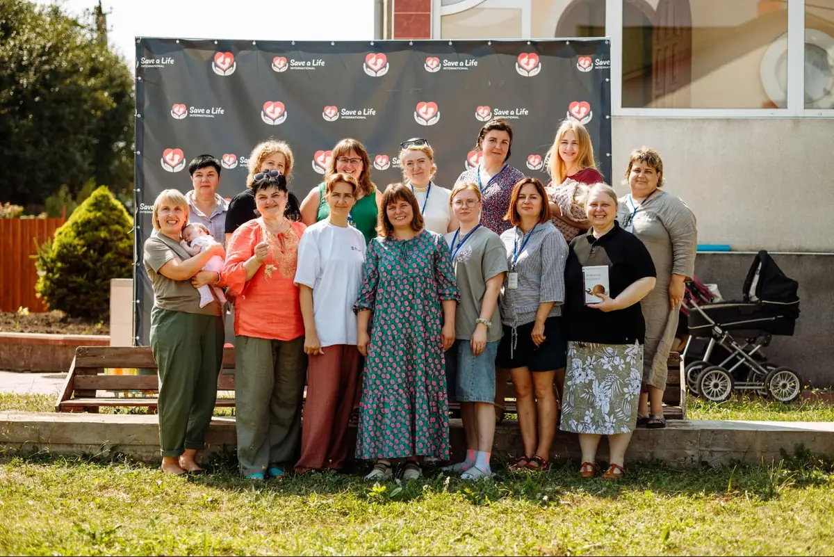 Mothers, some holding their babies, gather for a group photo in front of a Save a Life International banner at the camp.