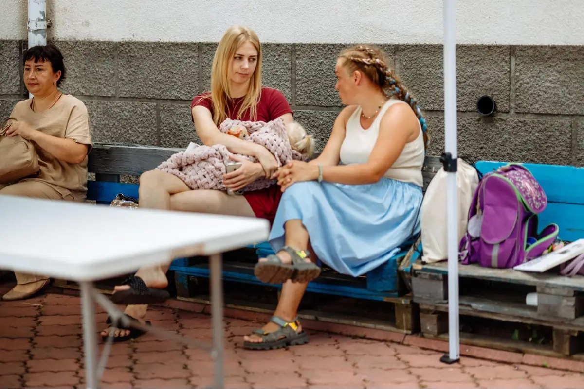 Mothers sit and talk on benches outdoors during a quiet moment at the “More Than Words” camp, one holding a sleeping child.